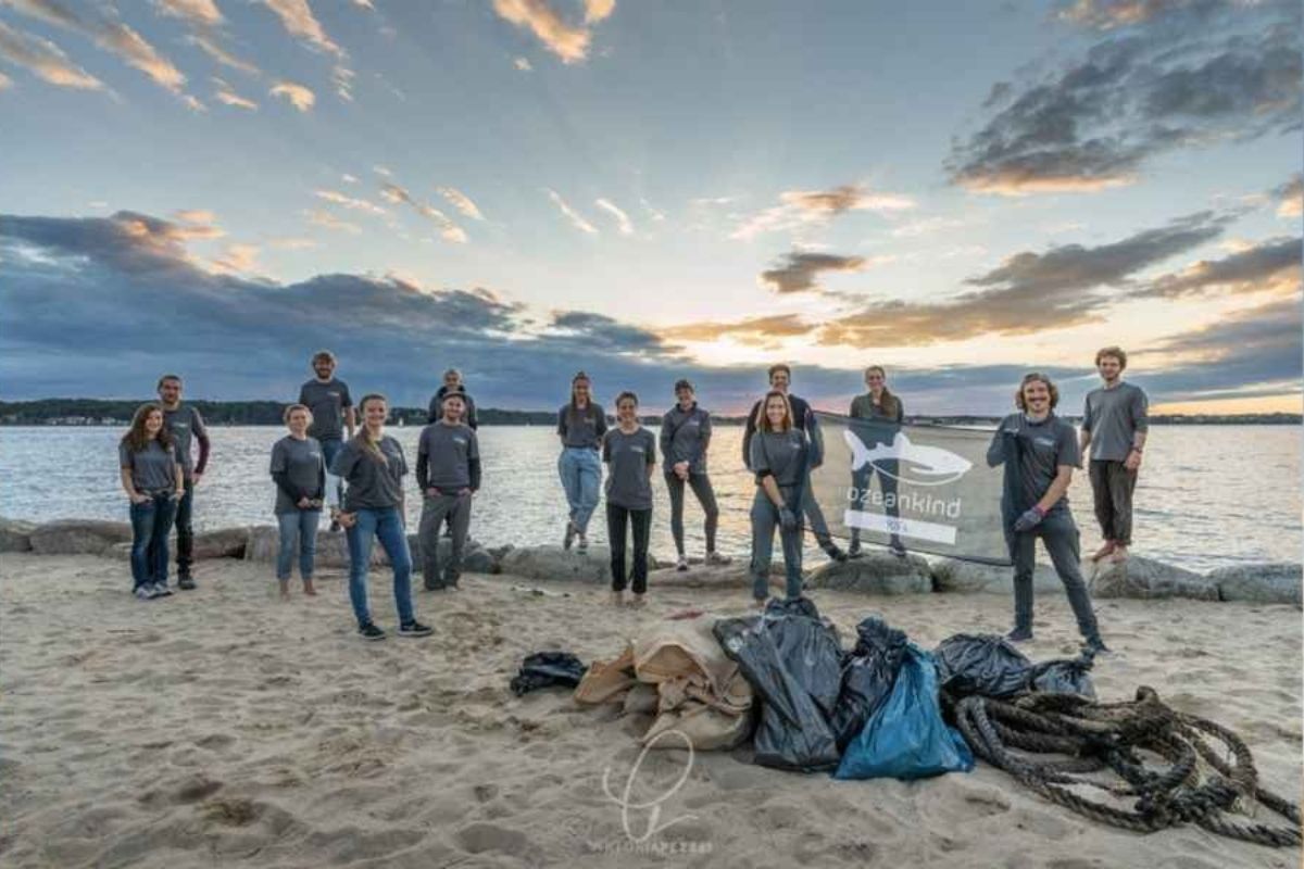 Mehrere Menschen stehen am Strand. Hinter ihnen ist das Meer zu sehen. Der Himmel ist blau, mit ein paar Wolken. Vor den Menschen liegen Mülltüten. Rechts sieht man zwei Menschen, die ein Schild von Ozeankind hochhalten.