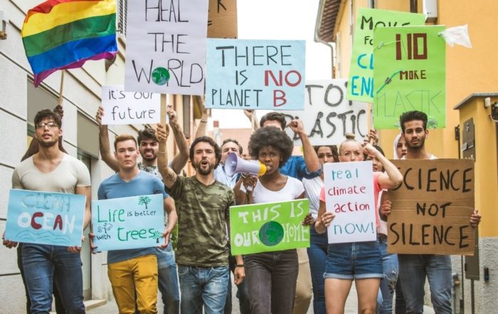 Auf dem Bild ist eine protestierende Menge zu sehen. Das Mädchen in der Mitte hat ein Megafon in der Hand und spricht darein. In der anderen Hand ist ein Schild mit dem Text: This is our home. Ganz rechts ist eine Regenbogenflagge zu sehen. Die texte auf den Schildern lauten von links nach rechts: Clean Ocean, Life is better with trees, it'sour future, heal the world, there is no Planet B, no more plastic, real climate action now, science not silence. Im Hintergrund sind Häuser zu sehen.