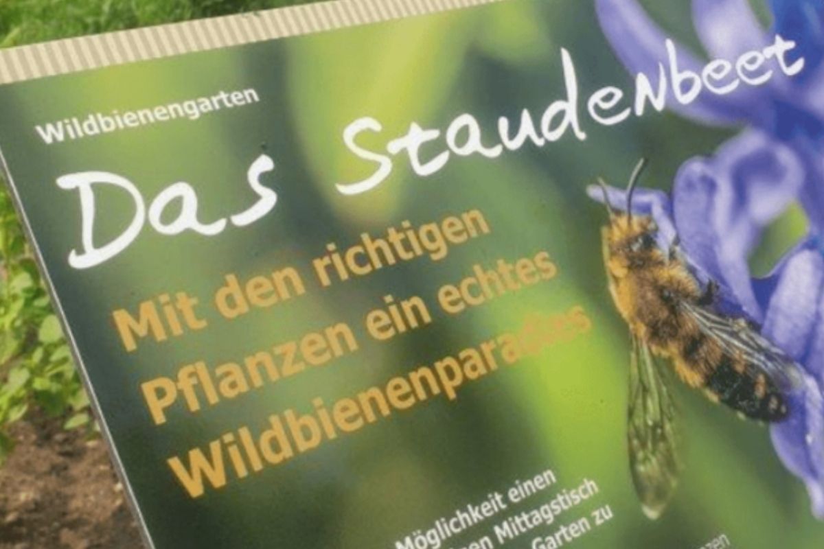 Ein grünes Schild mit der Abbildung einer Biene auf einer lila Blume am rechten Bildrand. Links zentriert steht in weißer und gelber Schrift "Das Staudenbeet. Mit den richtigen Pflanzen ein echtes Wildbienenparadies."