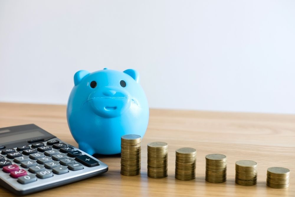 Das Foto zeigt einen Taschenrechner, ein blaues Sparschwein sowie sechs ordentliche Stapel mit Münzen. Es soll symbolisieren, dass es wichtig ist, sein Geld für nachhaltig agierende Unternehmen auszugeben und sinnvoll zu konsumieren.