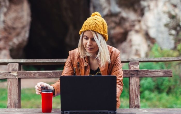 Eine blonde Frau sitzt in der Natur an einem Laptop. Im Hintergrund ist ein Park zu sehen. Sie hat einen Thermobecher in der Hand und schaut in ihr Gerät. Sie leistet ihren Beitrag im online Engagement im Naturschutz