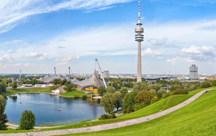 Es ist der Olympiapark in München zu sehen. Im Vordergrund ist die grüne Wiese, im Hintergrund die Skyline am Tag. Dieses Bild zeigt den Naturschutz in München.