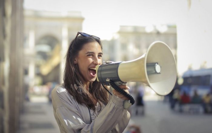 Eine junge Frau steht in einer Stadt. Sie hat ein Megaphon in der Hand und leistet Öffentlichkeitsarbeit im Naturschutz.
