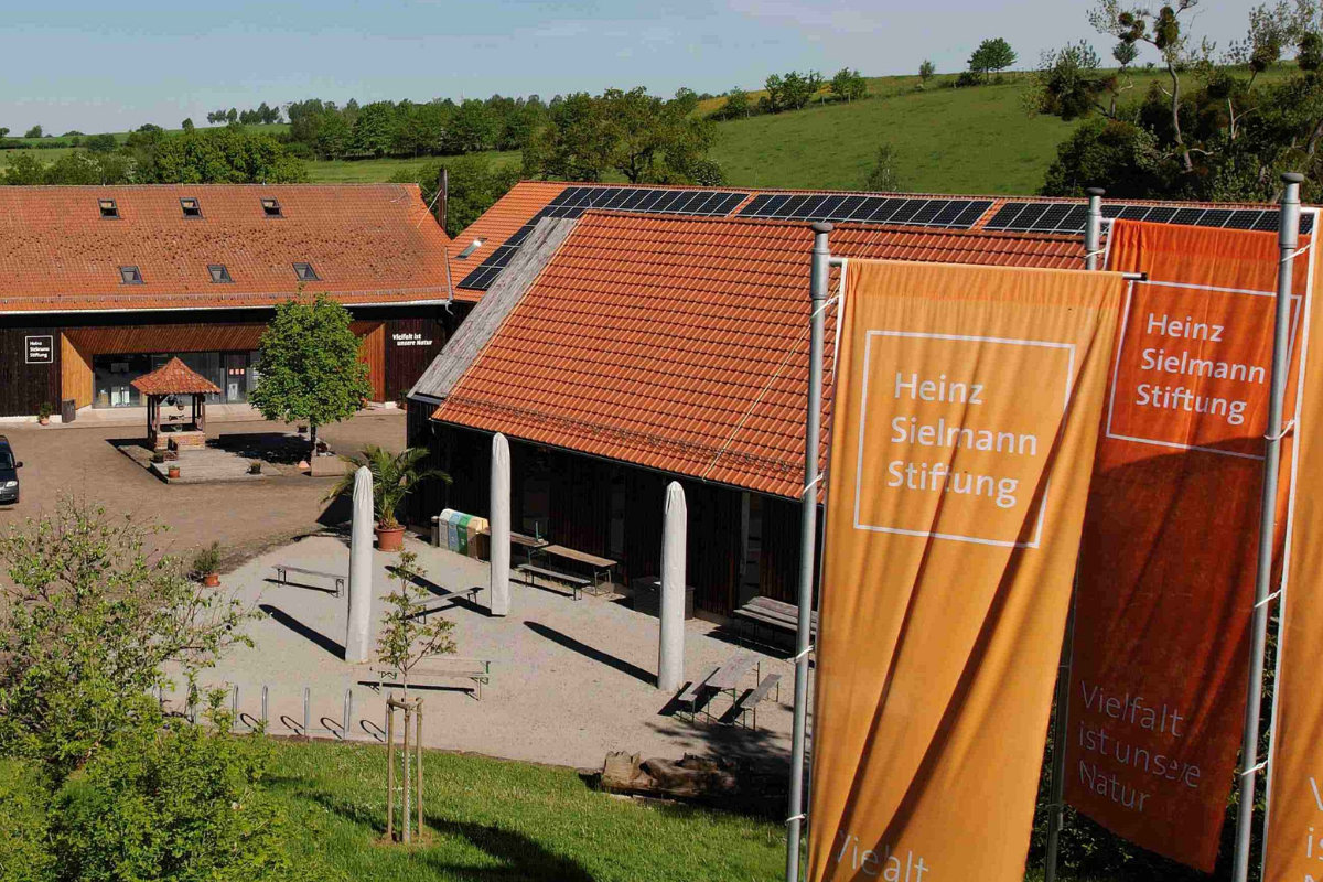 Das Foto zeigt das Gut Herbigshagen bei Duderstadt, einem alten Hof mit großen Scheunengebäuden und Ziegeldächern. Im Vordergrund wehen drei orangefarbene Fahnen mit dem Aufdruck "Heinz Sielmann Stiftung".