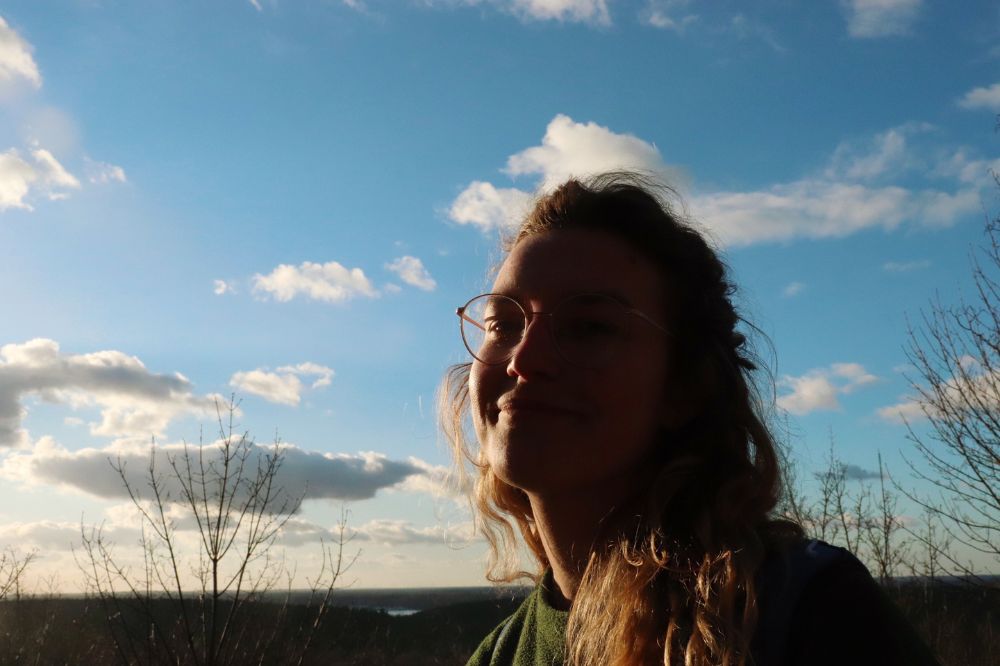 Auf dem Foto ist Marlene zu sehen. Sie lacht in die Kamera. Eine Hälfte ihres Gesichts wird von der Sonne angestrahlt. Im Hintergrund ist der blaue Himmel mit weißen Wolken zu sehen.